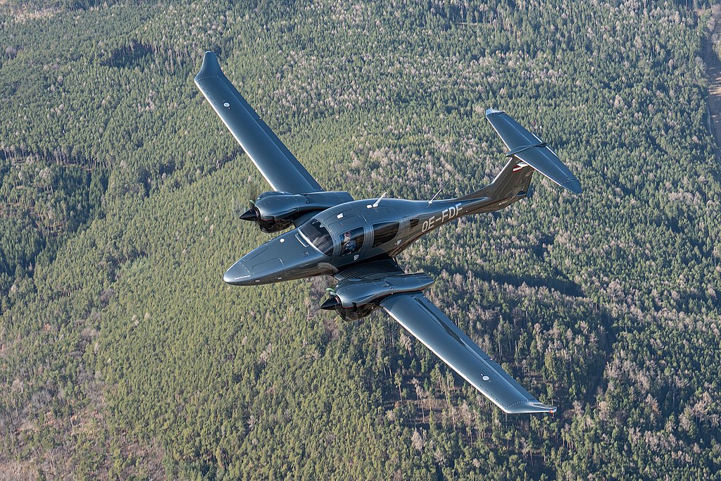 A DA62 aircraft glides through the sky, showcasing its shiny exterior against a backdrop of lush green forests below.