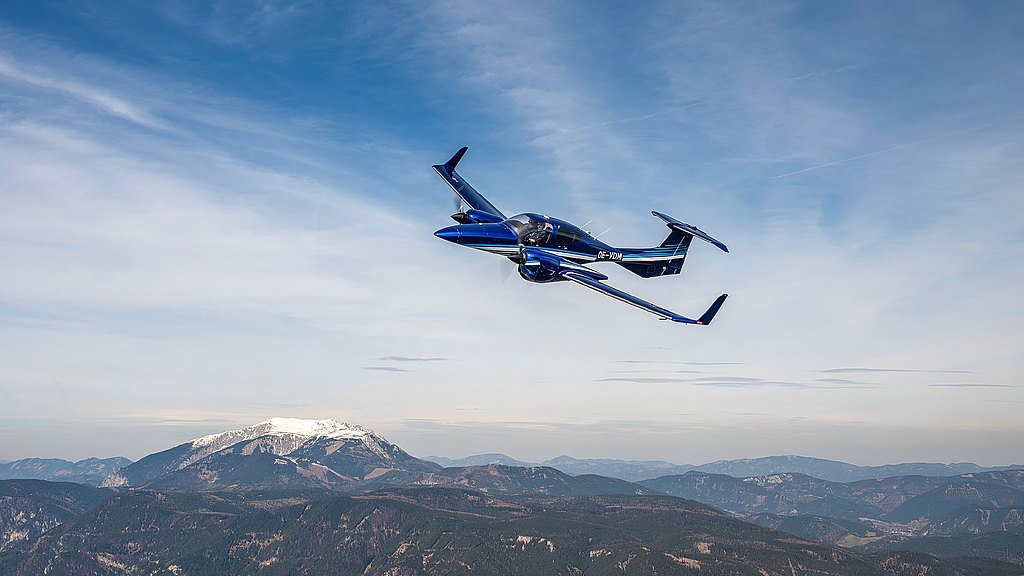 A sleek blue DA42-VI aircraft soars gracefully above a mountainous landscape, showcasing its modern design against a clear sky.