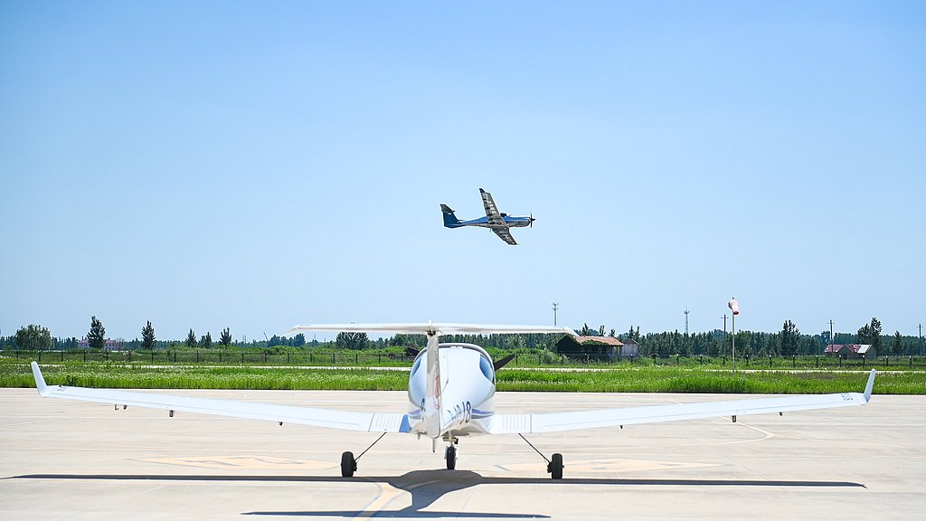 A DA40 aircraft is parked on a runway, while a DA50 RG flies overhead against a clear blue sky.