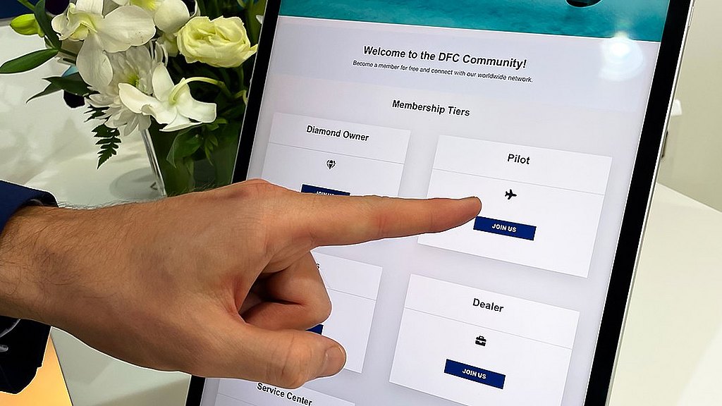A hand points at a tablet screen displaying membership options for the DFC Community, with flowers in the background.