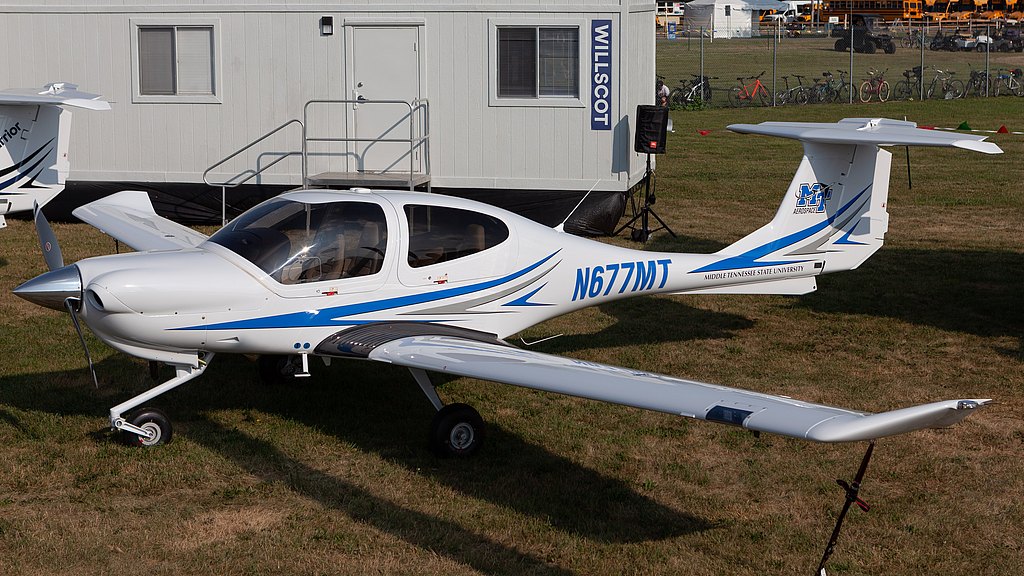 The image features a small airplane with a propeller, positioned on a grassy area near a portable building labeled 'WILLSCOT'.