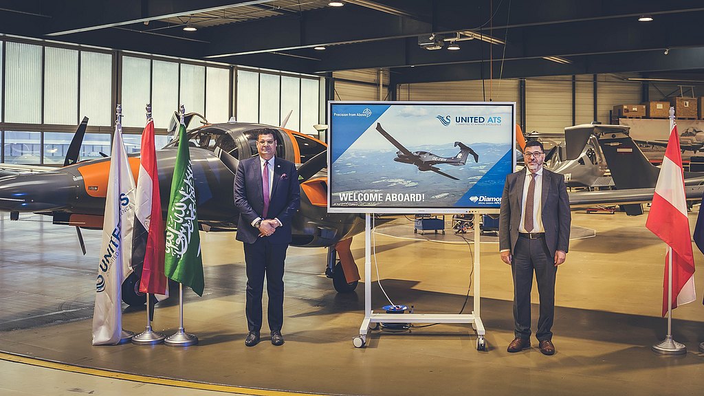Two men standing in an aircraft hangar next to a screen with a DA62 MPP in the back. 