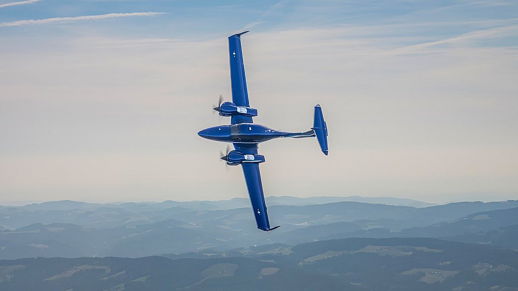 The blue DA42-VI elegantly banks in mid-air, showcasing its aerodynamic design against a backdrop of soft, layered hills.