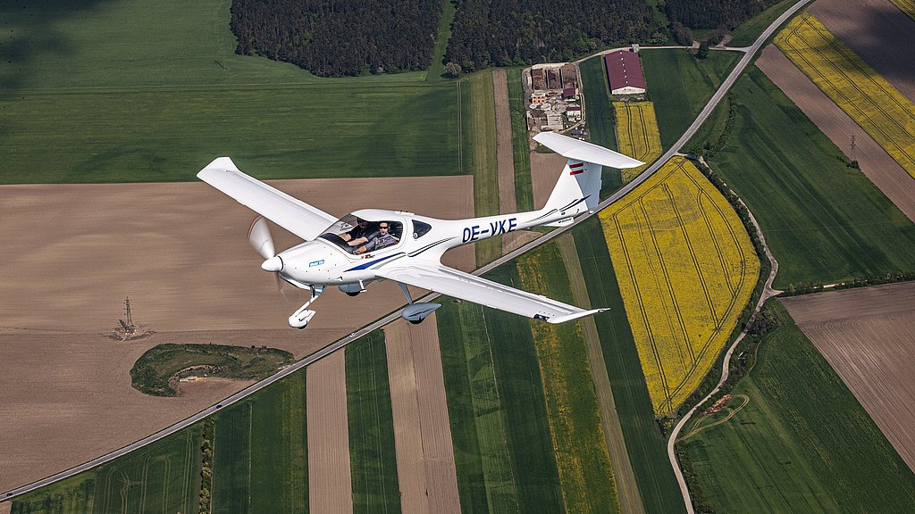 A DA20i Katana aircraft flies over vibrant green and yellow fields, showcasing the beauty of rural landscapes from above.