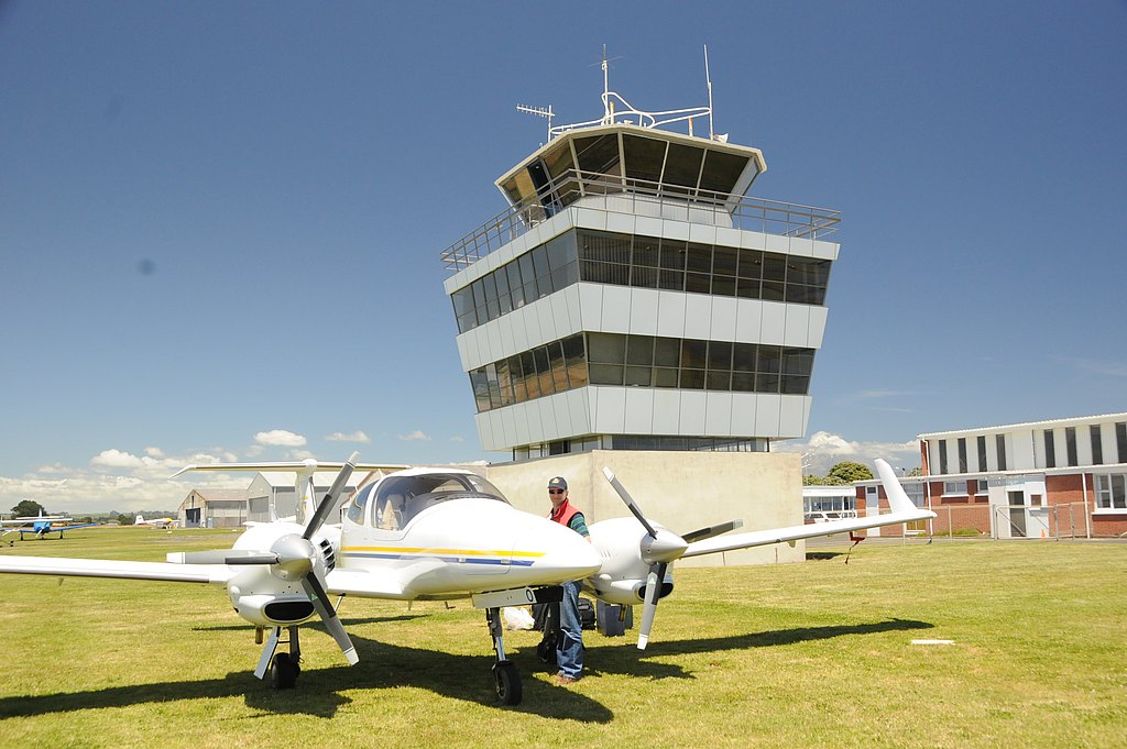 A DA42 aircraft stands on green grass beside a modern air traffic control tower under a clear blue sky.