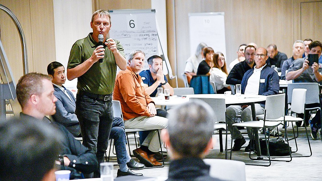 Man with a microphone talking to the participants while they are sitting on round tables.