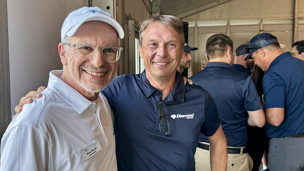 Two smiling men pose together, one wearing glasses and a white shirt, the other in a dark blue shirt with a logo.
