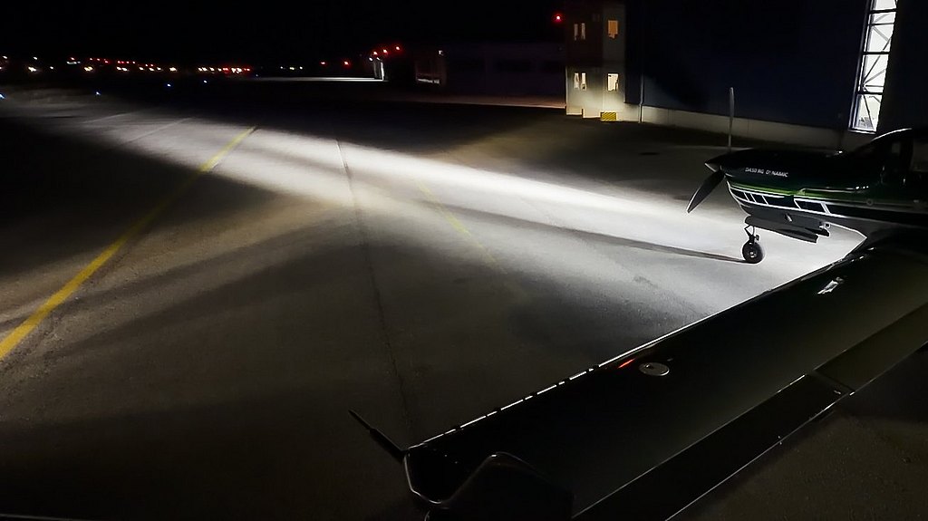 The image features a DA50 RG aircraft on a runway at night, with powerful LED taxi and landing lights illuminating the area and creating dramatic contrasts.