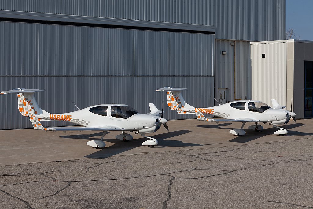 Two white DA40 NG aircraft with orange and gray patterns are parked on a concrete surface beside a large hangar.