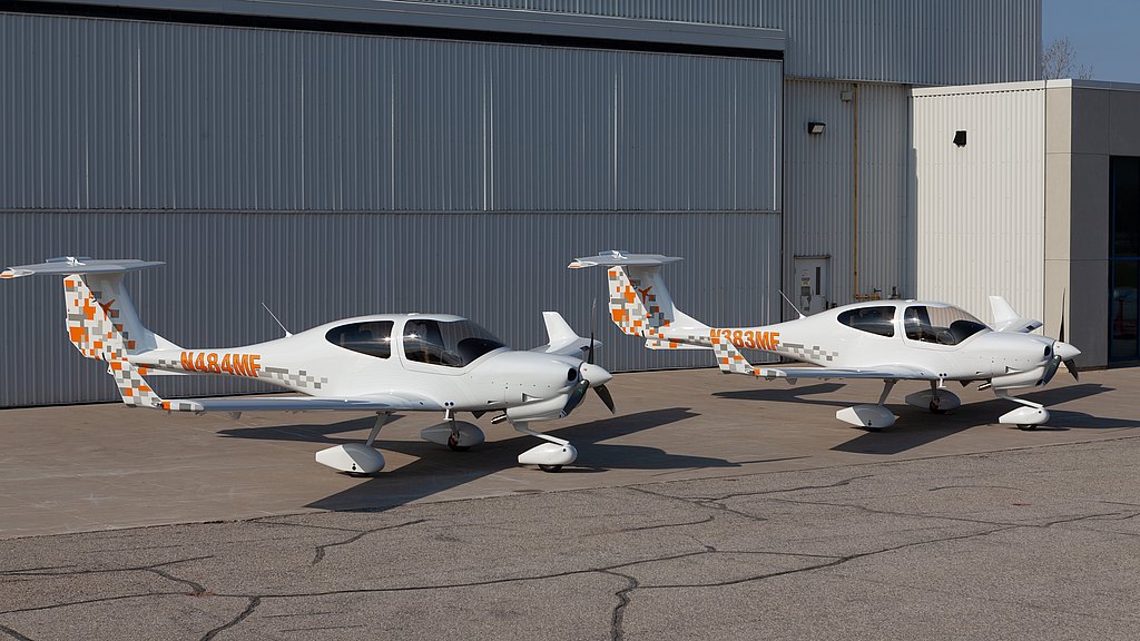 Two white DA40 NG aircraft with orange and gray patterns are parked on a concrete surface beside a large hangar.