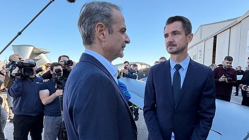 Amidst a gathering of people, two men face each other, deep in discussion, with a backdrop of aircraft and media presence.