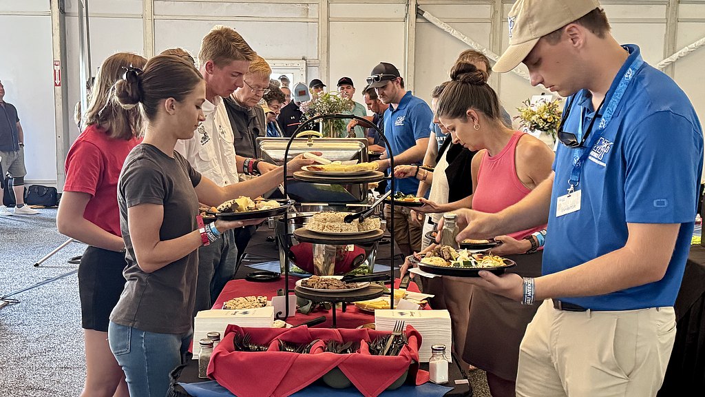 A group of people serve themselves from a buffet, selecting various dishes while enjoying a communal meal in a large tent.