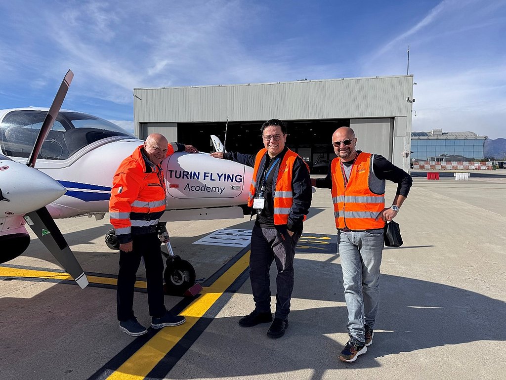 With a hangar in the background, three men in safety vests stand proudly by a DA42-VI training aircraft.