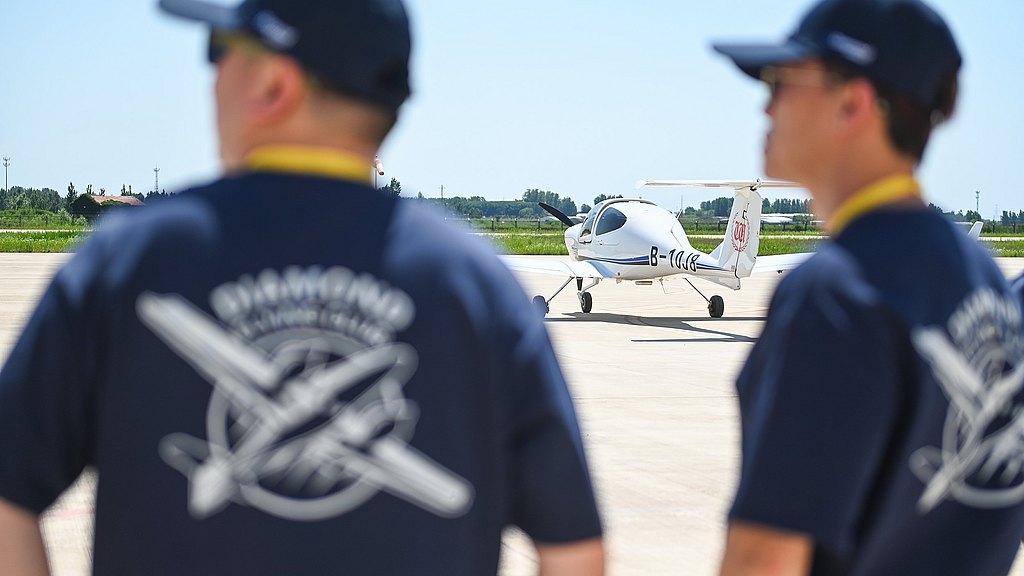 Two individuals in navy shirts observe a small aircraft preparing for takeoff on a sunny day at an airfield.