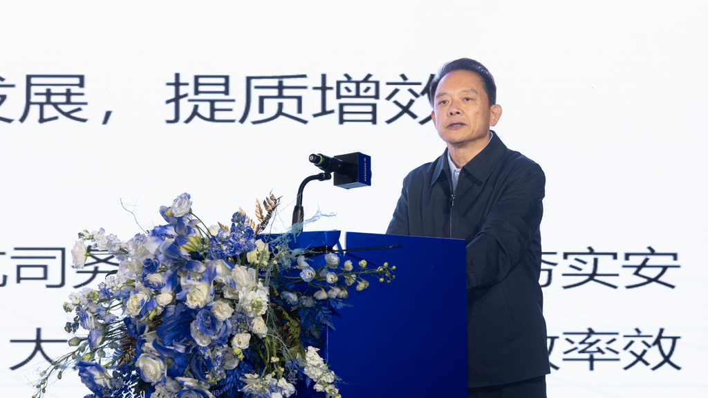In a formal setting, a man delivers a speech at a podium, surrounded by elegant floral arrangements and a bright backdrop.
