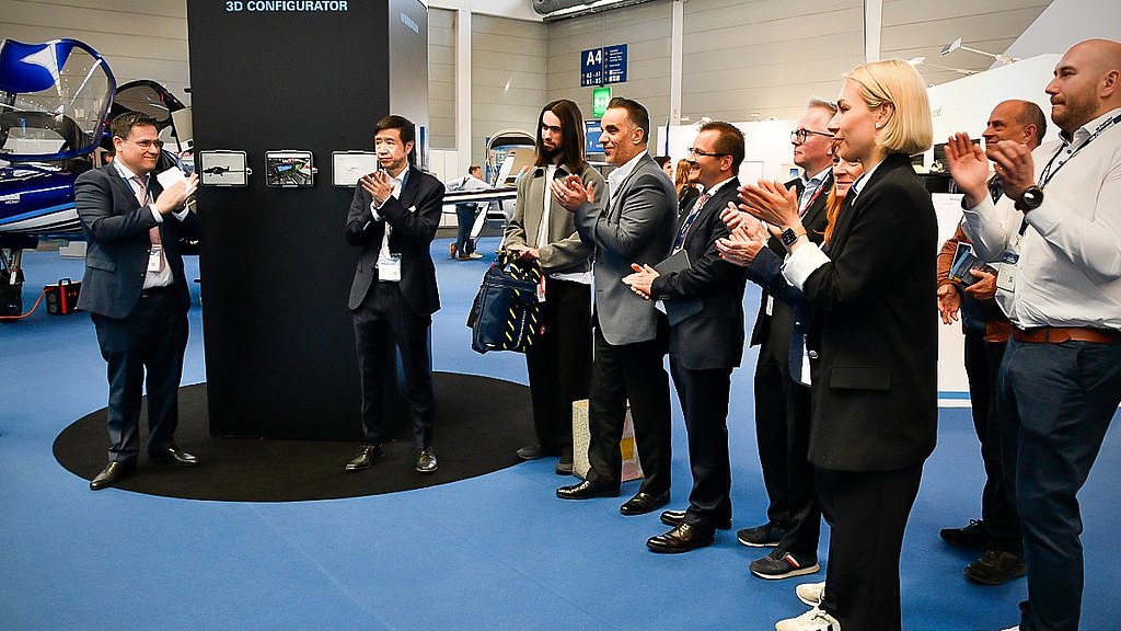 A group of professionals enthusiastically applauds a speaker presenting a 3D configurator at an aviation event.