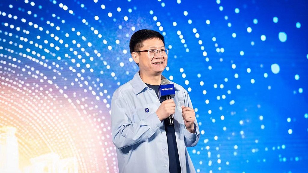 A man in a light blue shirt smiles while holding a microphone, speaking confidently against a vibrant, colorful backdrop.