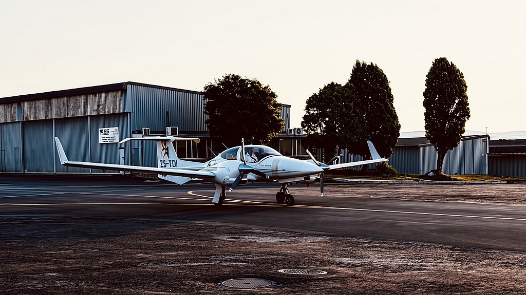 A white DA42 aircraft is parked on a tarmac with a well-lit hangar and trees visible in the background during sunset.