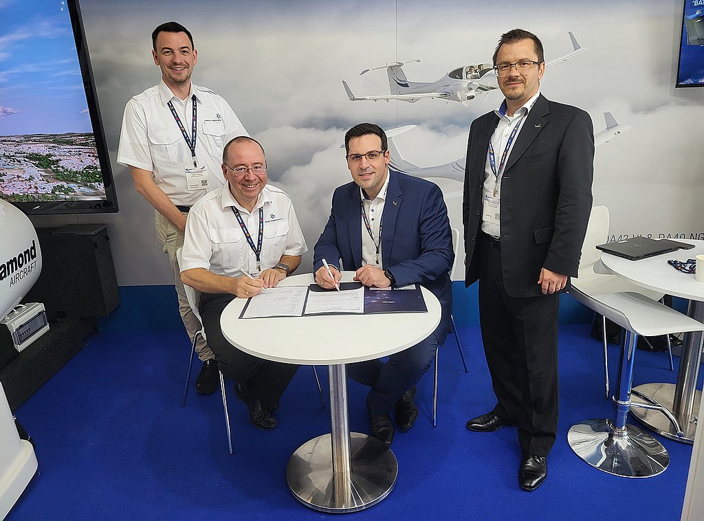 Four men sitting and standing around a coffee table at an event booth signing a contract.