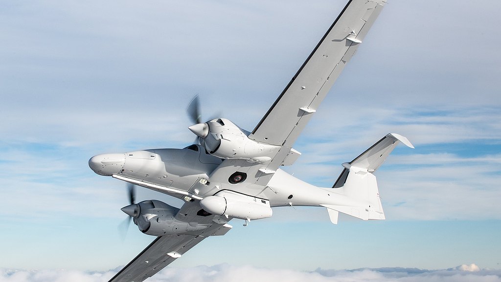The aircraft, painted in white, banks sharply against a backdrop of fluffy clouds and a bright, expansive sky.