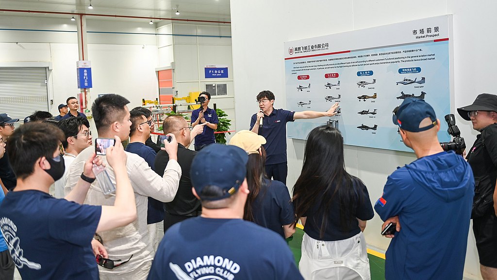 A presenter explains various aircraft models to an engaged audience in a well-lit industrial setting, with a display board behind.