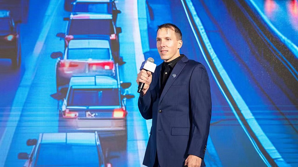 A man in a blue suit stands confidently on stage, holding a microphone, with a backdrop of busy traffic.