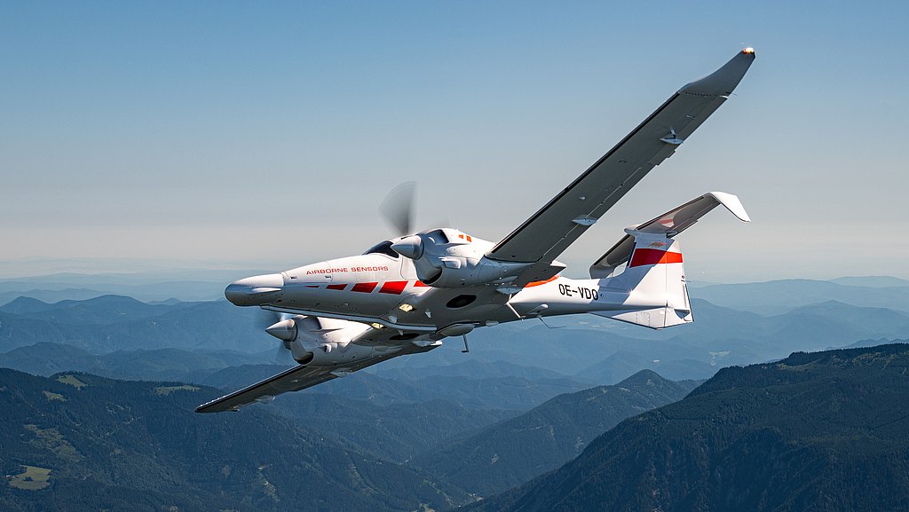 A DA62 MPP aircraft with a sleek design flies over a mountainous landscape, showcasing its distinctive markings.
