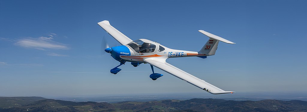 A blue and white DA20i Katana aircraft soars above lush green hills under a clear blue sky, showcasing its elegant design.