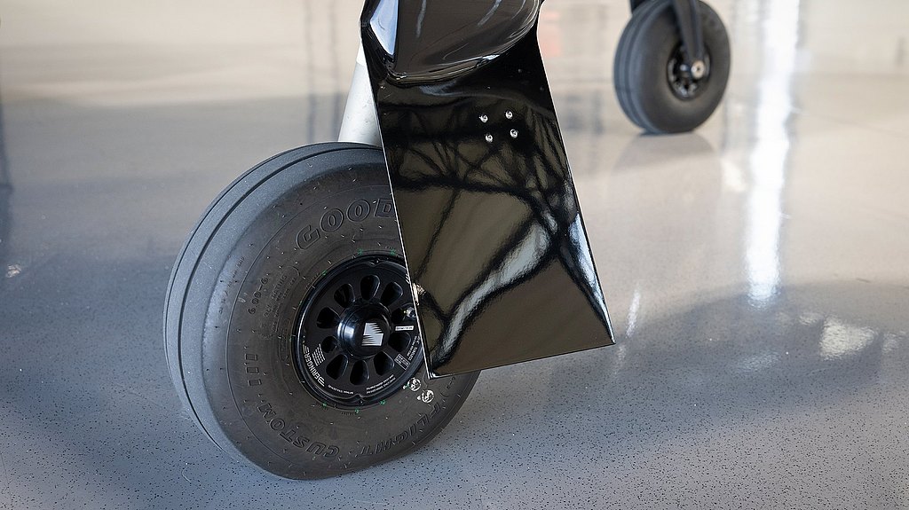 The image highlights a robust aircraft tire mounted on a black wheel, reflecting light against a smooth, glossy floor.