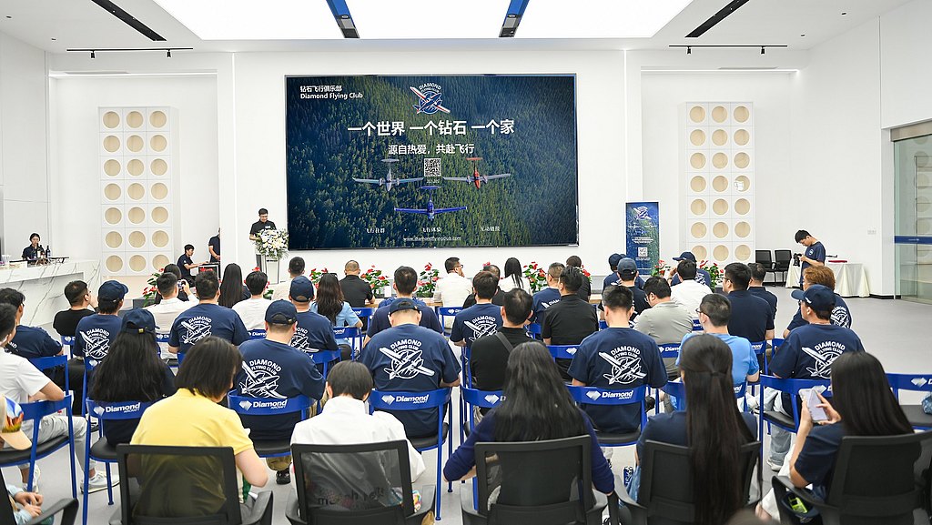 A group of attendees in blue shirts sits in a modern conference room, focused on a speaker at the front.