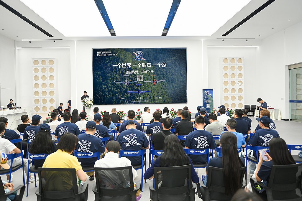 A group of attendees in blue shirts sits in a modern conference room, focused on a speaker at the front.