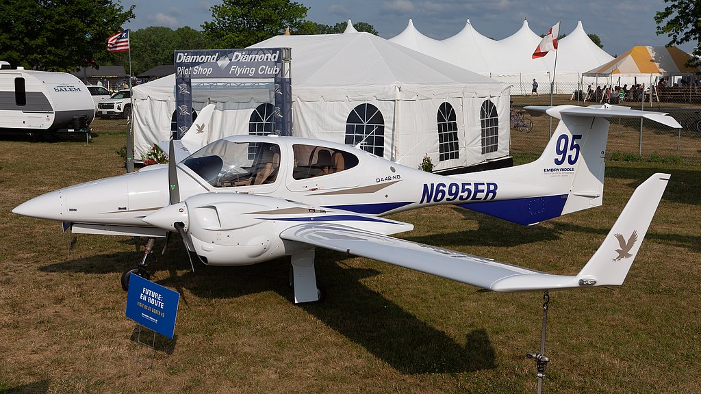 The Diamond DA42 aircraft stands prominently, with a sign indicating its future route, set against a lively outdoor backdrop.