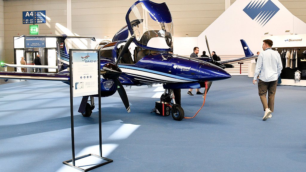 A blue DA42-VI aircraft with an open canopy is showcased at an aviation exhibition, surrounded by attendees exploring the venue.