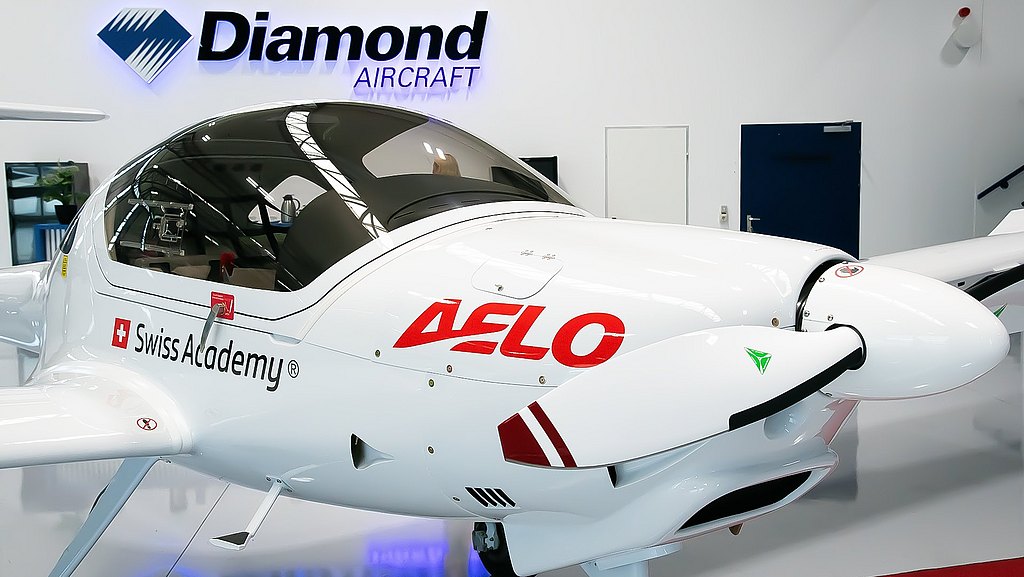 A white DA20i Katana aircraft with the logo 'AELO' prominently displayed, parked inside a modern aviation facility.