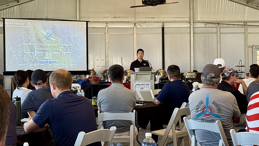 A speaker presents to an audience in a large tent, with a projector displaying a flying club's logo and message.