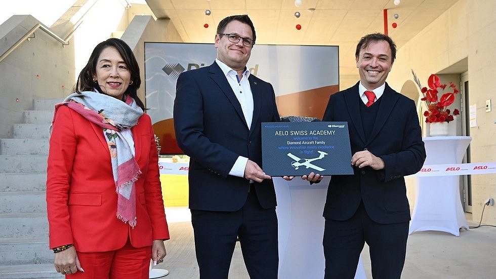 In a modern setting, a woman in red and two men in suits proudly present a plaque for the Aelo Swiss Academy.
