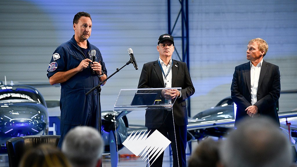 Man in a flight suit speaking with a microphone on the stage. 