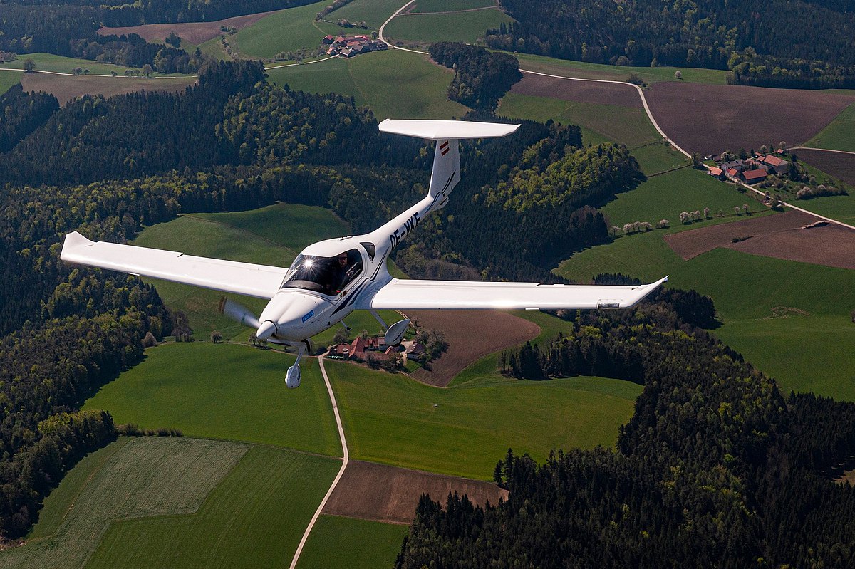 A DA20i Katana aircraft glides gracefully over lush green fields and dense forests, showcasing the beauty of the landscape below.