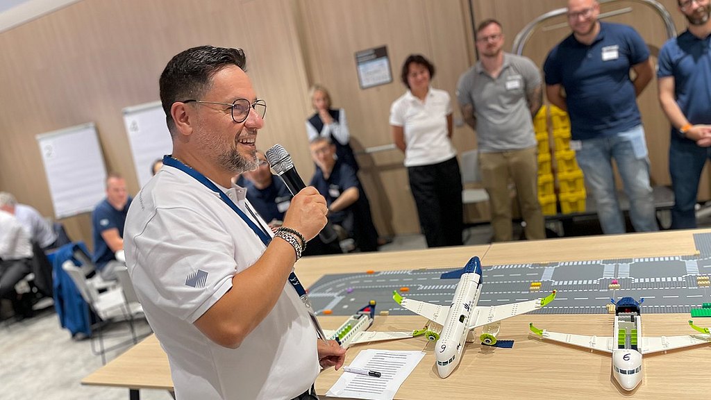 Man with a microphone talking to the participants of the workshop while standing next to the finished Lego aircraft.