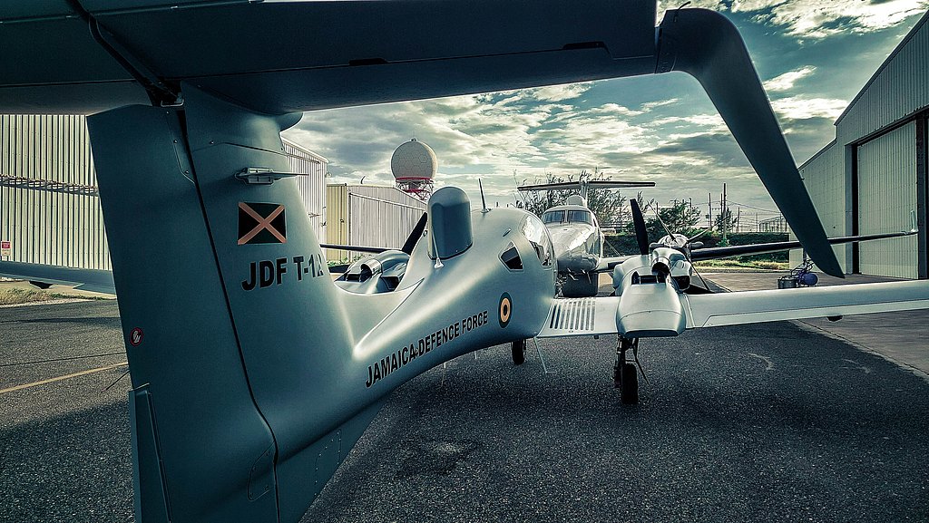 A DA62 MPP aircraft with 'Jamaica Defence Force' markings is parked on a tarmac, surrounded by hangars and a cloudy sky.