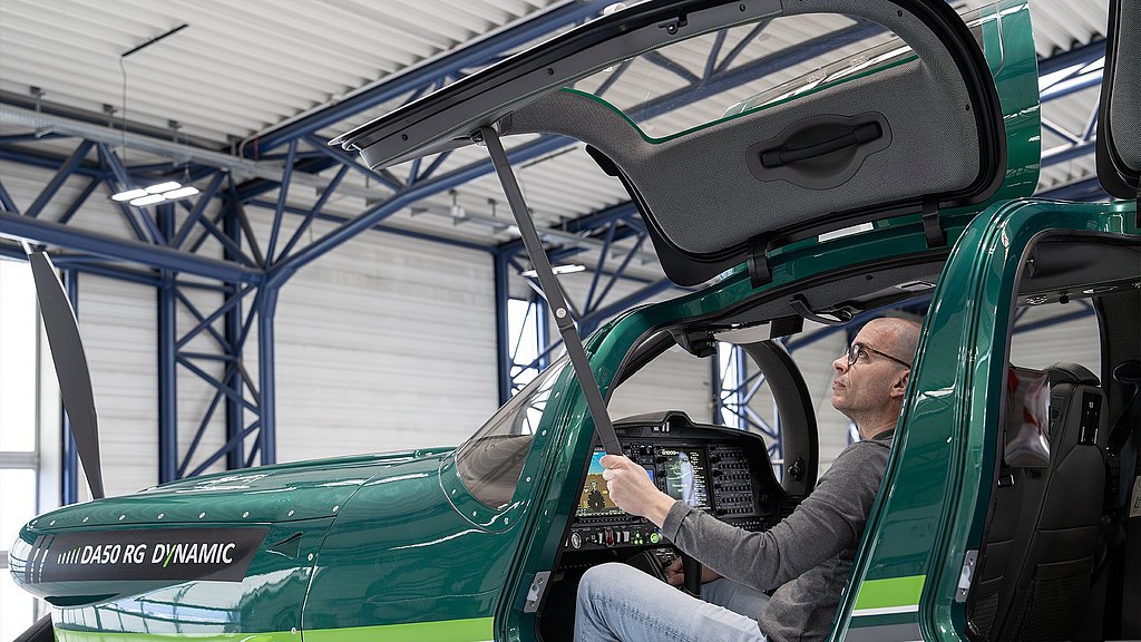 In a bright hangar, a man pulls down the door closing strap of a DA50 RG aircraft to easily close the gullwing door. 