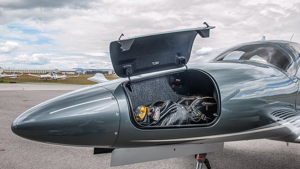 Inside the aircraft's nose compartment, a black bag and a yellow object are visible, framed by a dramatic sky and distant planes.