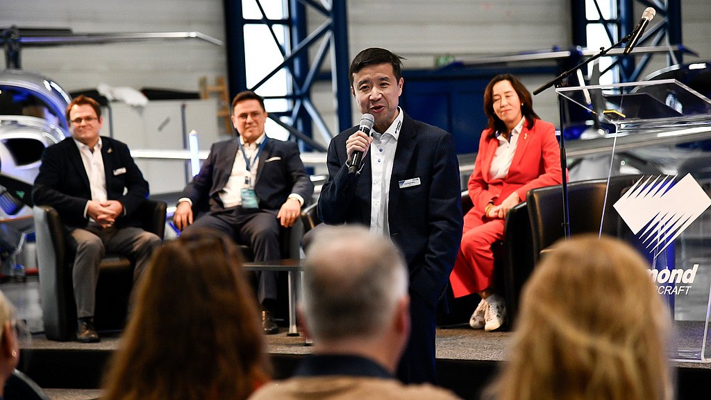 Man speaking to the audience with the stage and other panel discussion speakers in the back. 