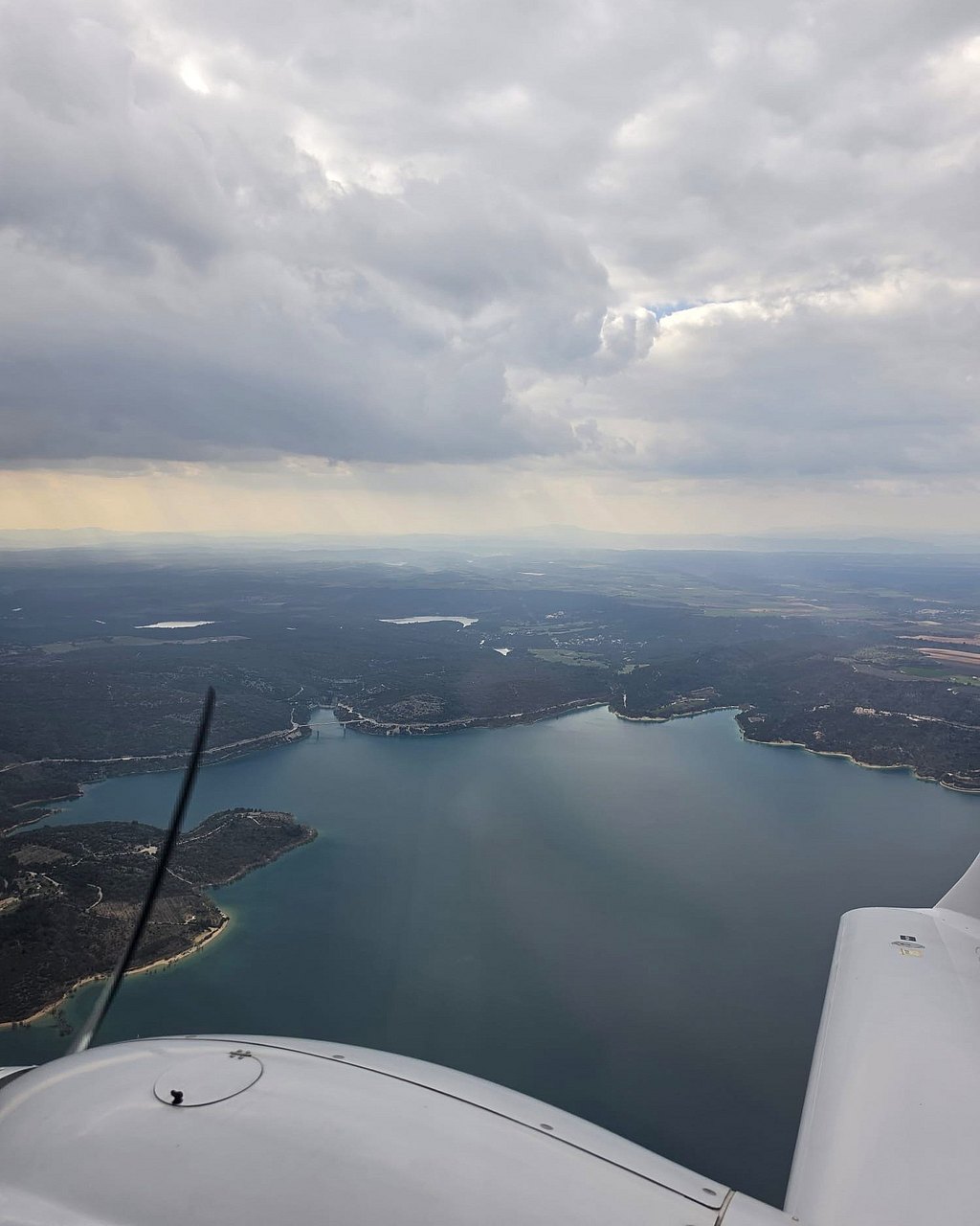 The landscape features a tranquil lake bordered by forests and fields, seen from an airplane wing against a dramatic sky.