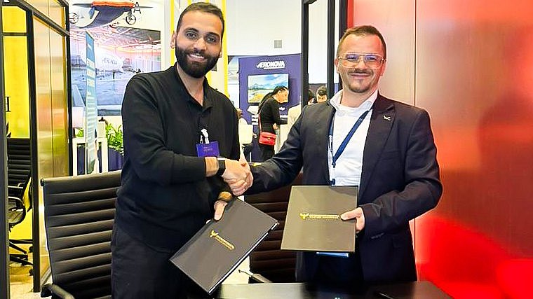 Two men shake hands in front of the Intercontinental Aviation Academy booth, holding signed documents and smiling confidently.