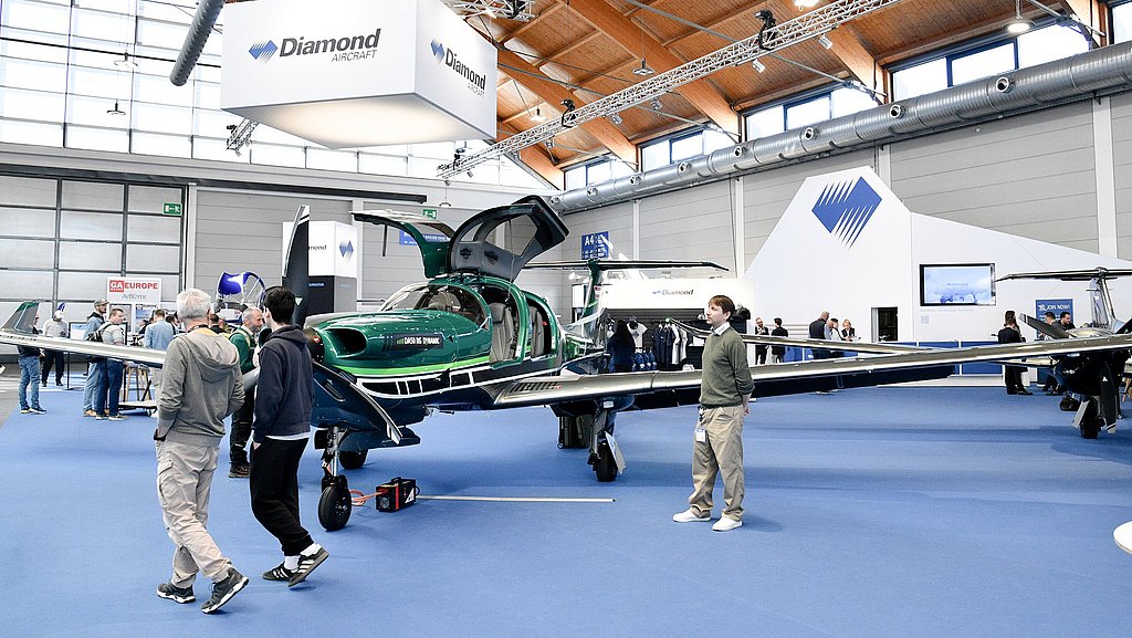 A green DA50 RG aircraft with its doors open is showcased at an aviation exhibition, surrounded by interested attendees.