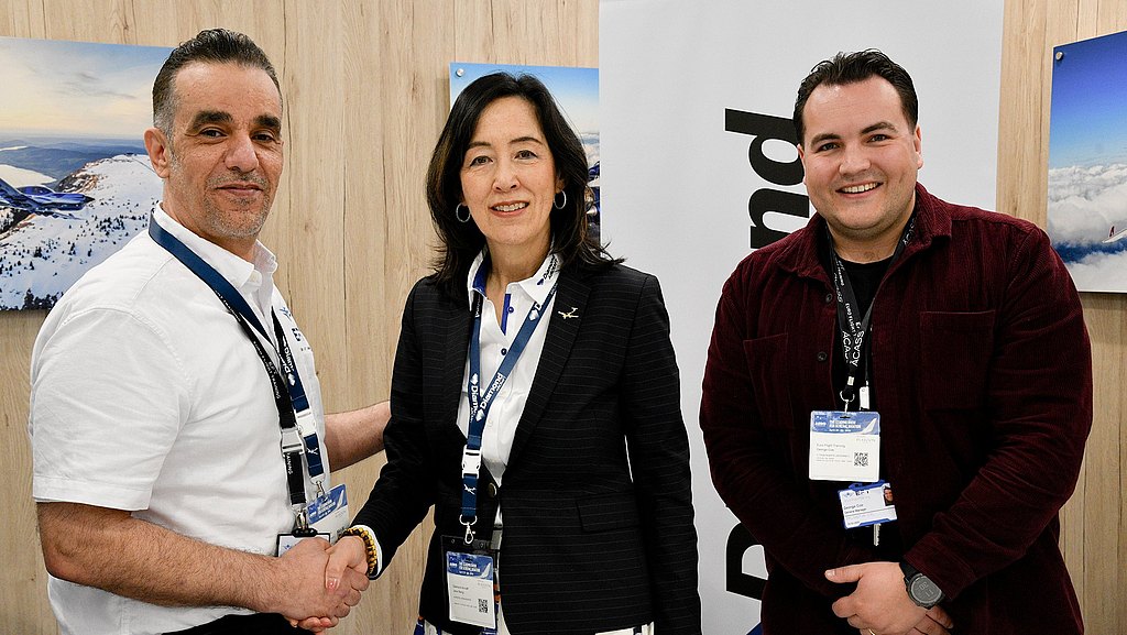 Three professionals engage in a handshake, showcasing collaboration at an aviation event, with a backdrop of scenic aviation images.