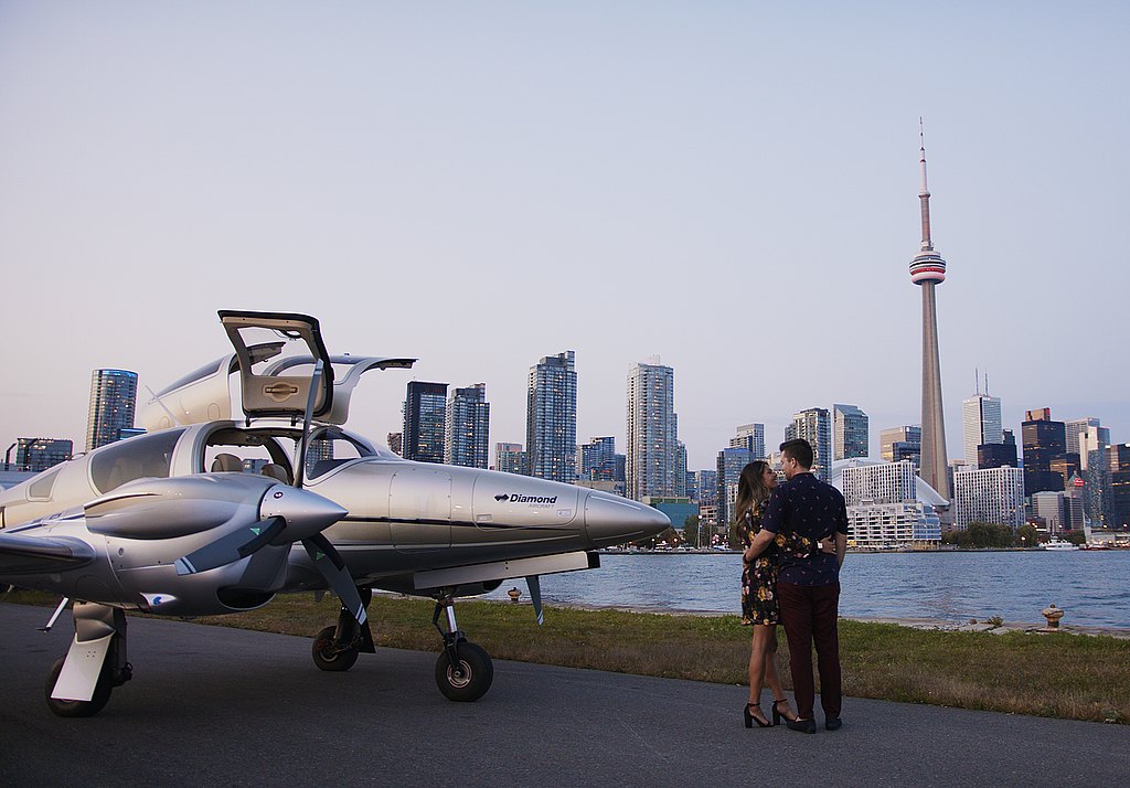 The couple stands close, gazing into each other's eyes, with a stylish DA62 plane and Toronto's iconic skyline as their backdrop.