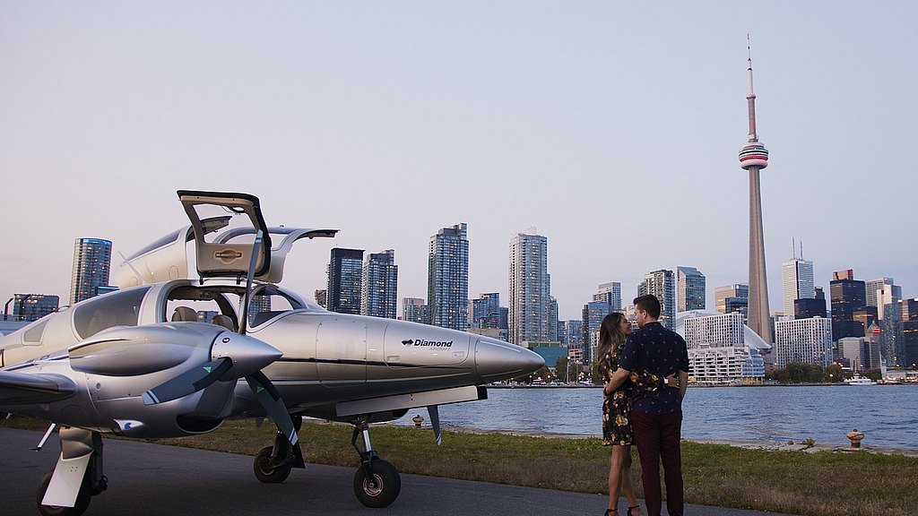 The couple stands close, gazing into each other's eyes, with a stylish DA62 plane and Toronto's iconic skyline as their backdrop.