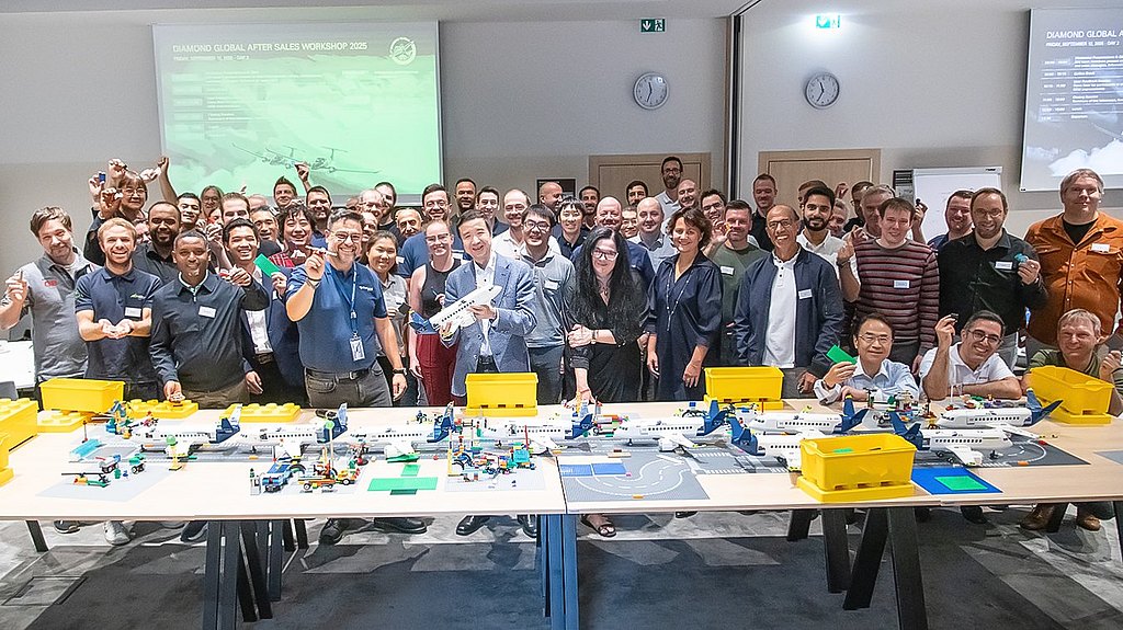 Large group of people standing behind a desk with Lego aircraft in a seminar room. 